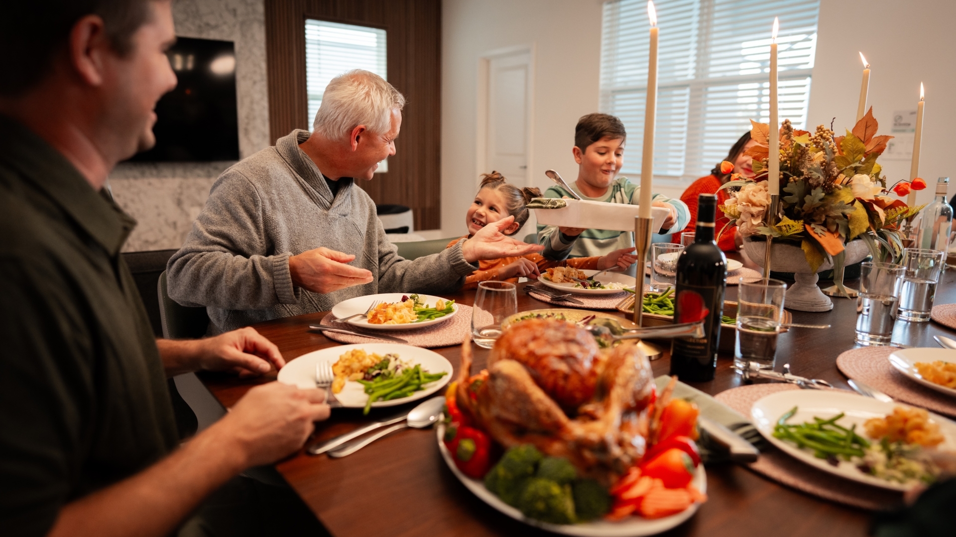 A multi-generational family sits around a large wooden dining table enjoying a holiday meal, featuring a roasted turkey platter in the foreground, lit taper candles, and an autumn floral centerpiece.