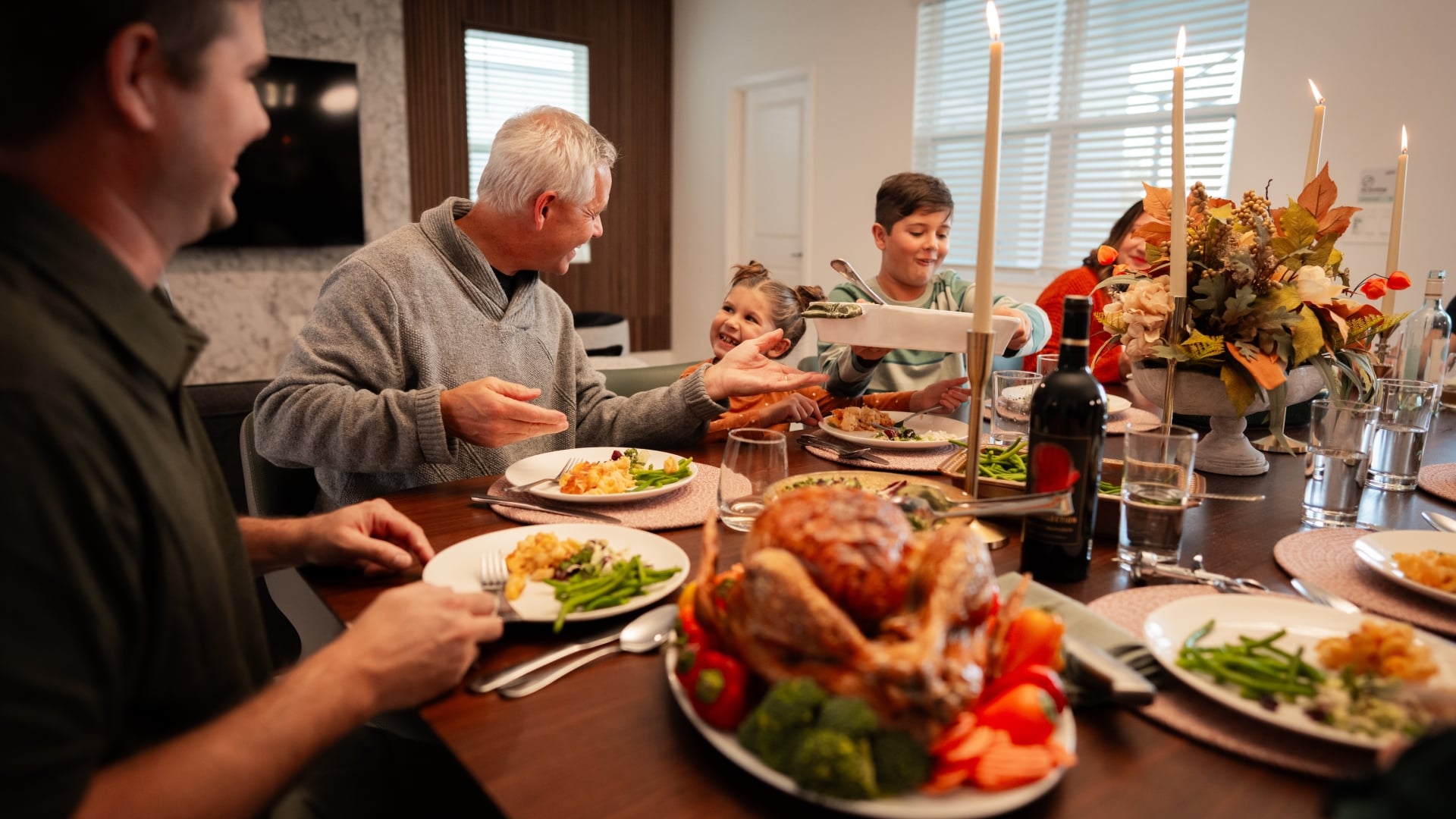 A multi-generational family sits around a large wooden dining table enjoying a holiday meal, featuring a roasted turkey platter in the foreground, lit taper candles, and an autumn floral centerpiece.