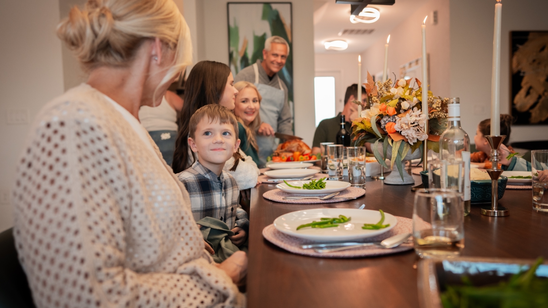 A multi-generational family sits around a large wooden dining table enjoying a holiday meal, featuring place settings, lit taper candles, and an autumn floral centerpiece.