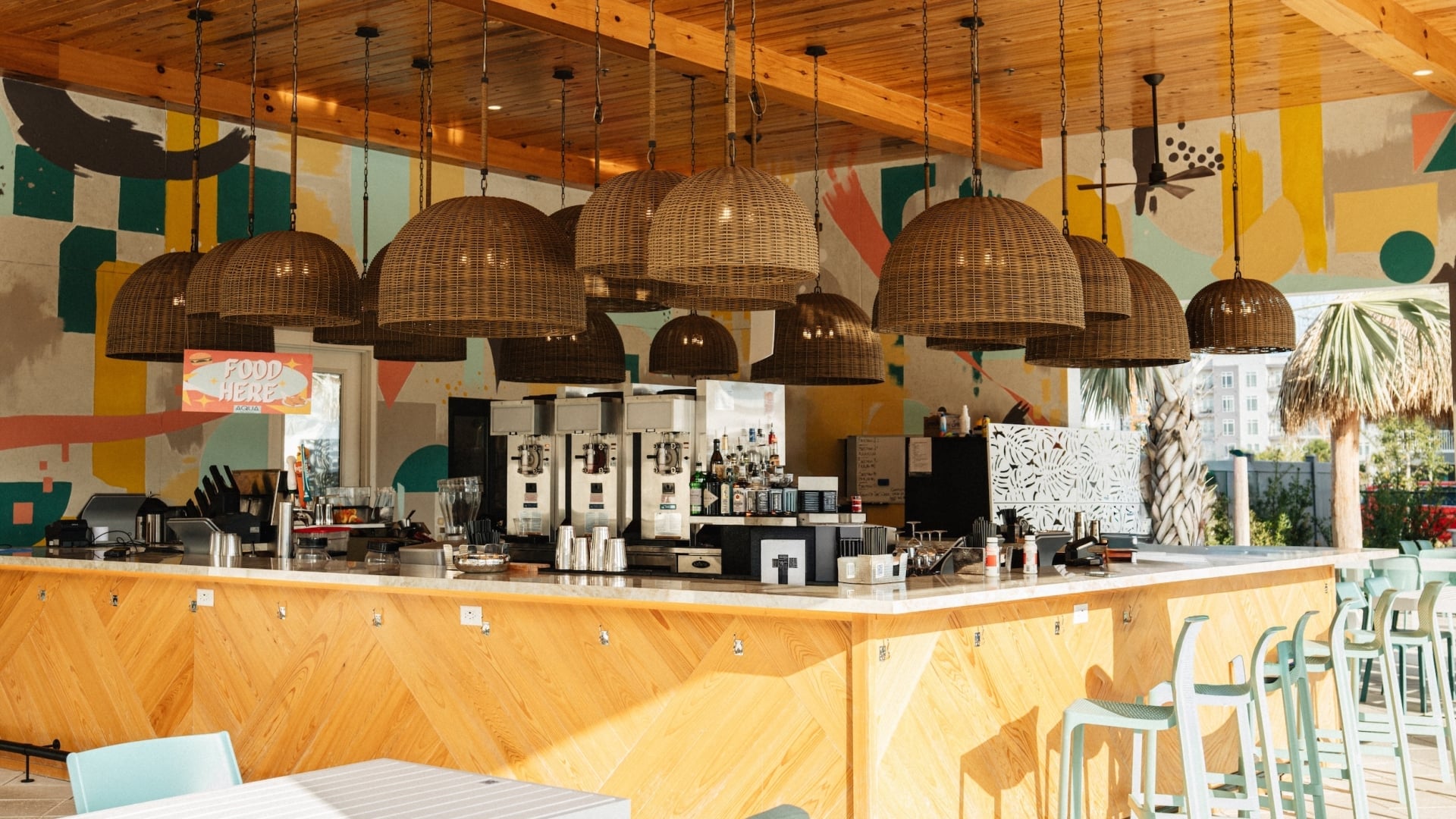 An open-air resort bar under a polished wood ceiling with woven basket pendant lights, featuring a light wood island counter, mint-green barstools, and a colorful graphic mural background.