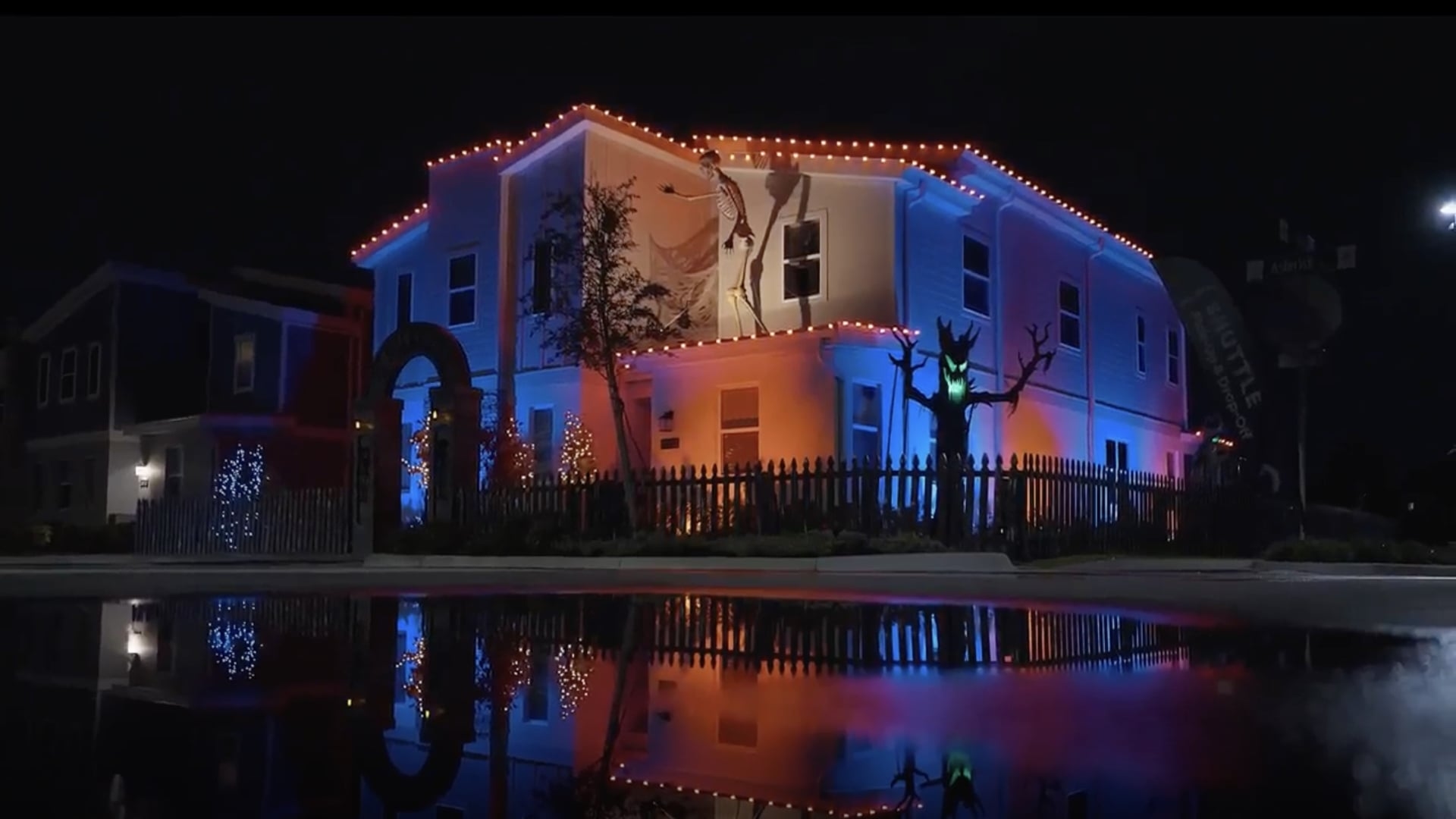 A two-story resort villa converted into a haunted house at night, illuminated by orange roofline lights and featuring a glowing green-eyed tree monster and a puddle reflection.