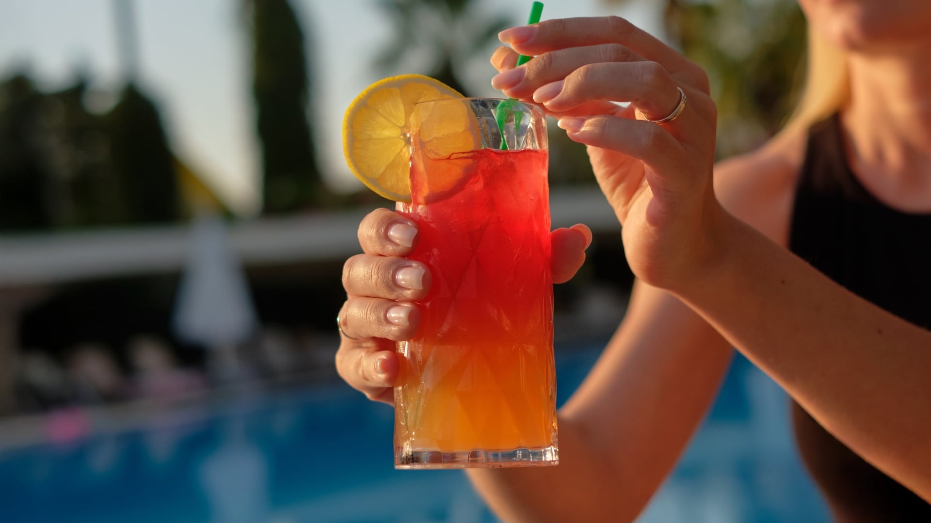 A close-up view of a woman's hands holding a layered red and orange tropical cocktail garnished with a lemon wheel and a green straw, set against a blurred poolside background.