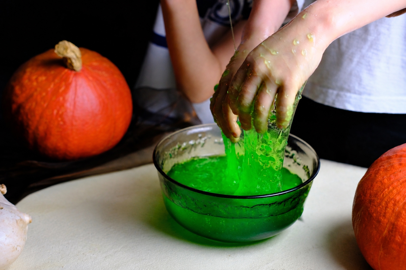 A close-up view of a child's hands mixing a bright green slime craft in a glass bowl, flanked by pumpkins on a table.