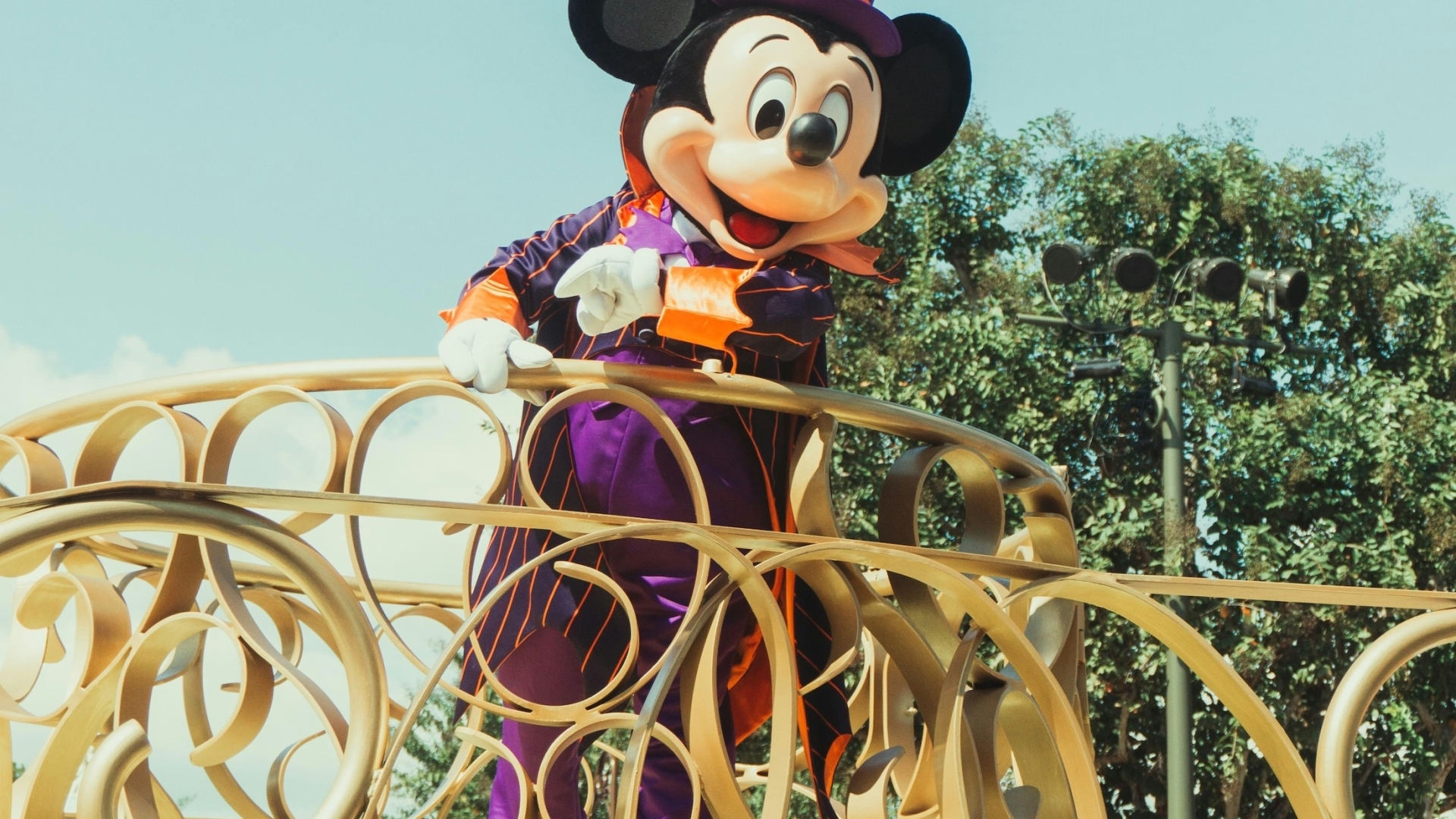Mickey Mouse wearing a purple and orange Halloween tuxedo with a matching top hat, waving from an ornate gold parade float balcony under a clear blue sky.